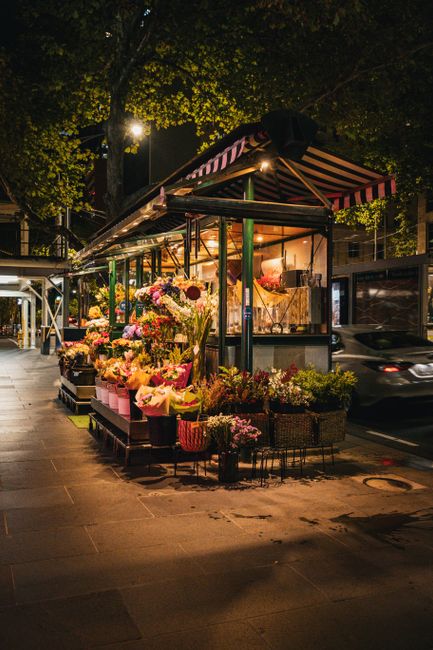 A nighttime scene of a flower stall on a city sidewalk, featuring a variety of fresh floral arrangements in vibrant colors. The display includes pink, purple, yellow, and white flowers, arranged in ba