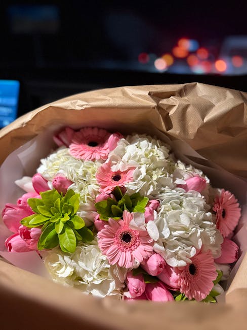 A floral arrangement featuring white hydrangeas and pink gerbera daisies, wrapped in light brown kraft paper with white tissue paper inside. The bouquet includes vibrant green foliage, and the flowers
