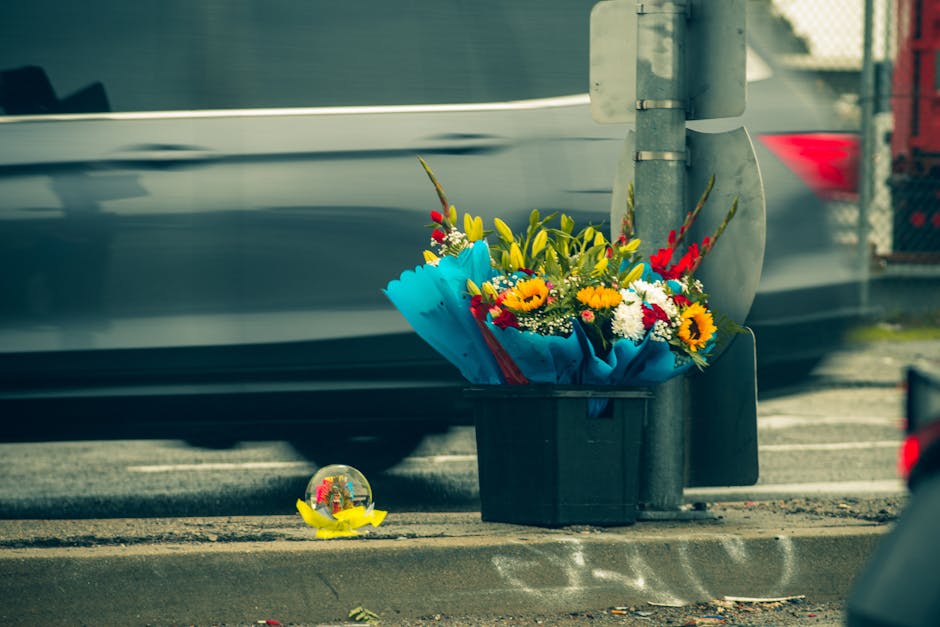 A colorful bouquet of fresh flowers including sunflowers, roses, and lilies, wrapped in bright blue paper and placed in a black container on the sidewalk near a streetlight. The arrangement features v