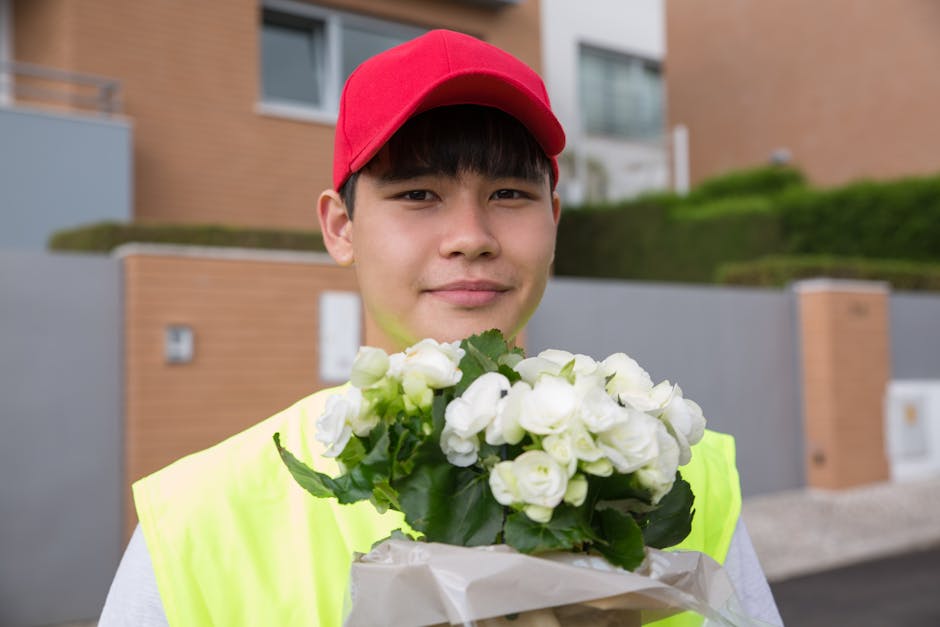 A young delivery person wearing a bright yellow high-visibility vest and a red cap is holding a bouquet of fresh white roses wrapped in clear plastic and white paper, with green leaves visible among t