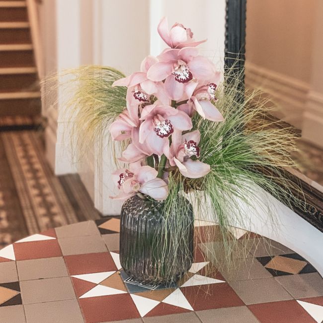 A tall floral arrangement featuring large, pale pink orchids with intricate, dark pink and white markings on their lips, set in a dark, textured glass vase. The orchids are complemented by soft, wispy