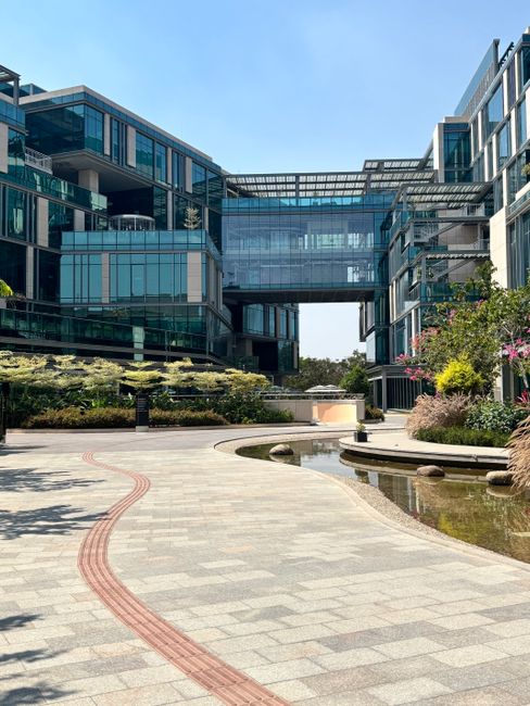 The image depicts a modern corporate office building with extensive glass facades reflecting the blue sky. In the foreground, a spacious, paved courtyard features a curved red brick pathway, neatly ar